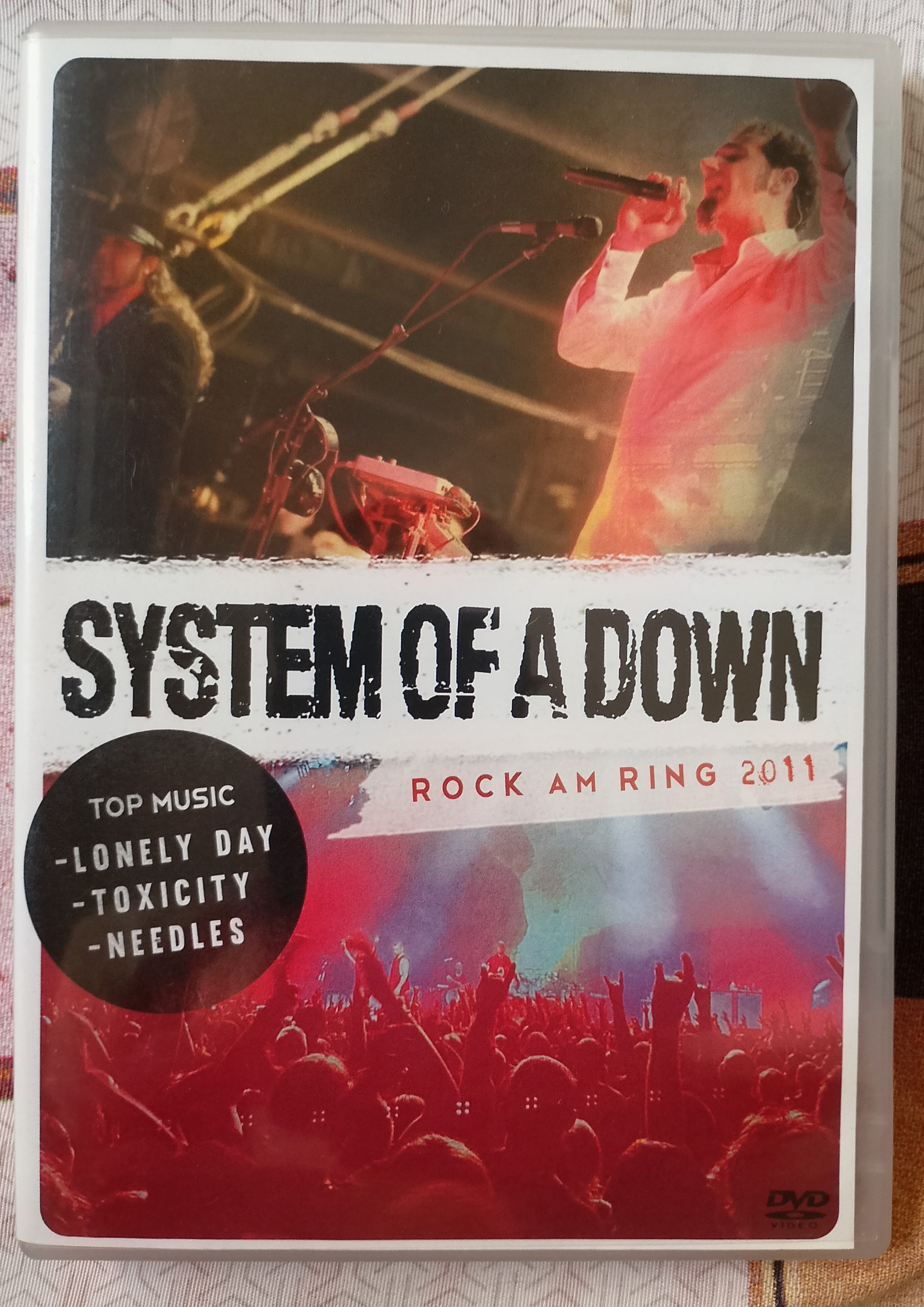 Capa de DVD de um show da banda System of a Down no Rock am Ring 2011, destacando as músicas Lonely Day, Toxicity e Needles.