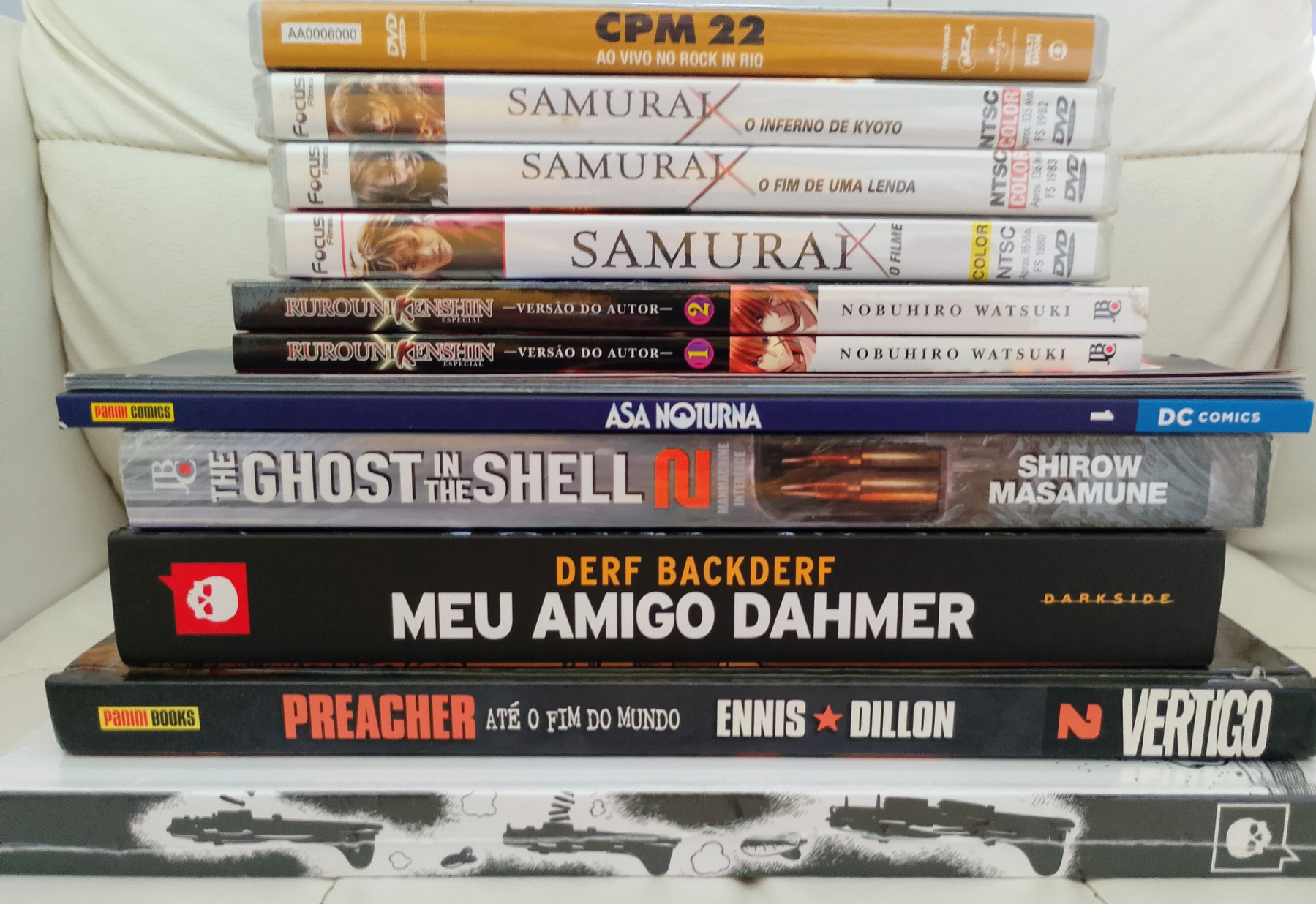 Auto-generated description: Há uma pilha de DVDs e livros, incluindo títulos como Samurai X, The Ghost in the Shell e Preacher.